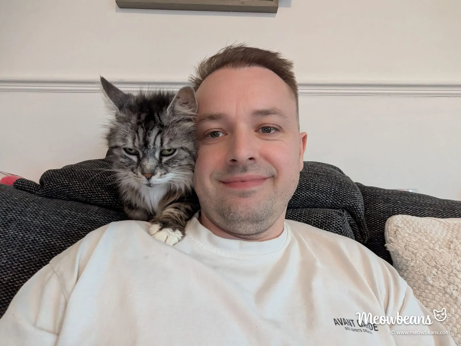John Maddock smiling on the sofa with Bean, his silver tabby Maine Coon, on his shoulder
