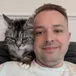 John Maddock smiling on the sofa with Bean, his silver tabby, on his shoulder