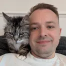 John Maddock smiling on the sofa with Bean, his silver tabby, on his shoulder